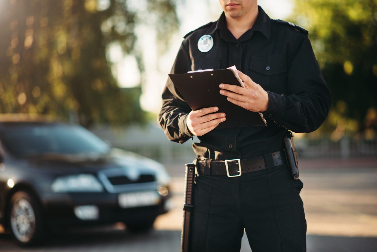 A police officer in a dark uniform holds a clipboard, standing by a blurred dark car outdoors.