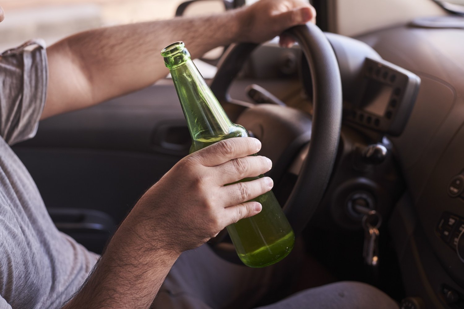 A person holds a green beer bottle while gripping a steering wheel