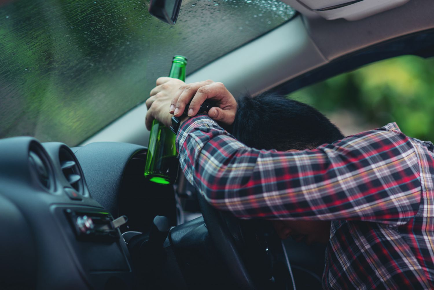 A person with a green bottle is slumped over a car's steering wheel