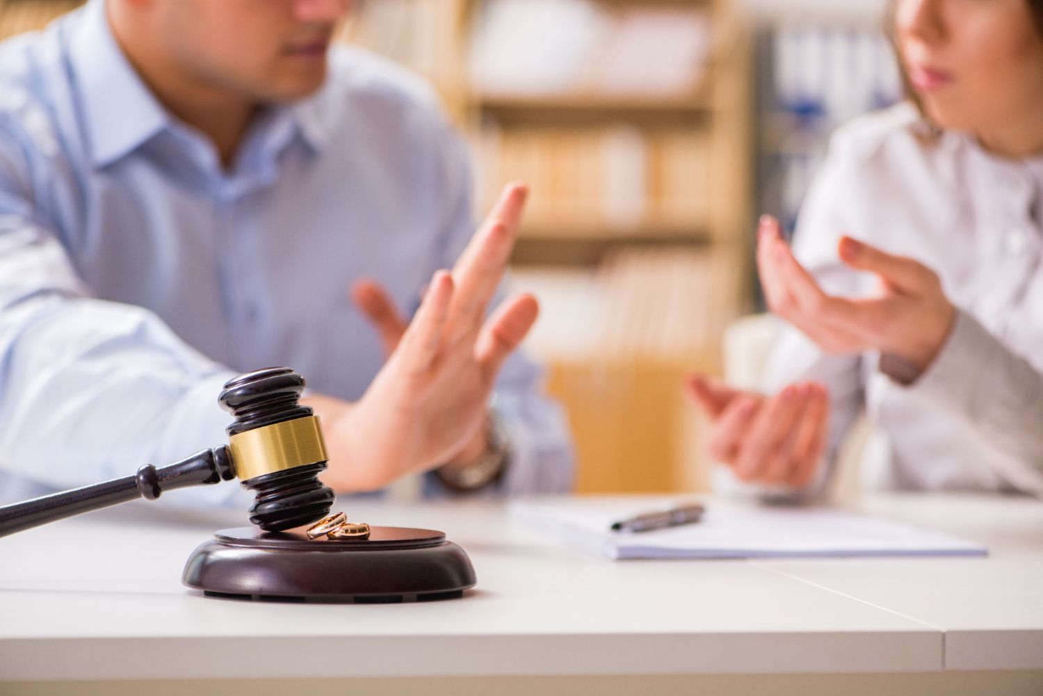 A gavel and wedding rings rest on a table as a couple engages in a serious discussion
