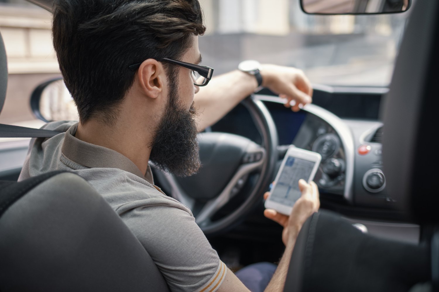 A bearded man wearing glasses uses his smartphone while seated in the driver's seat