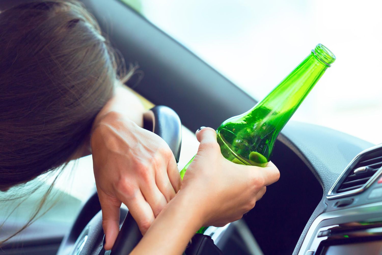 A person inside a car, holding a green glass bottle while their head rests on the steering wheel