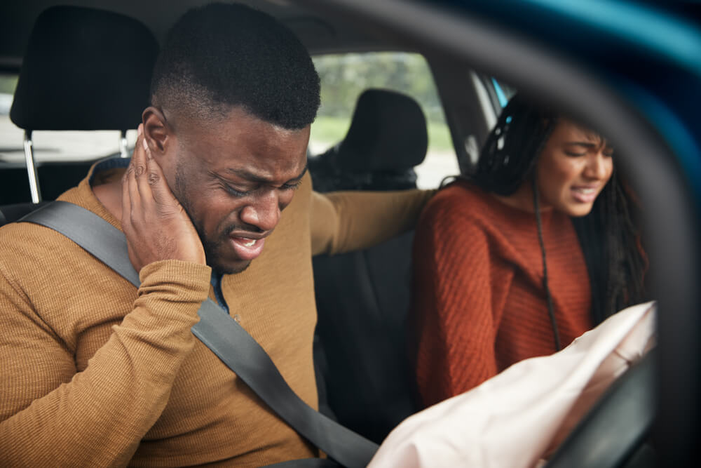 A man clutches his neck in pain after a car accident, while a distressed woman sits next to him with a deployed airbag