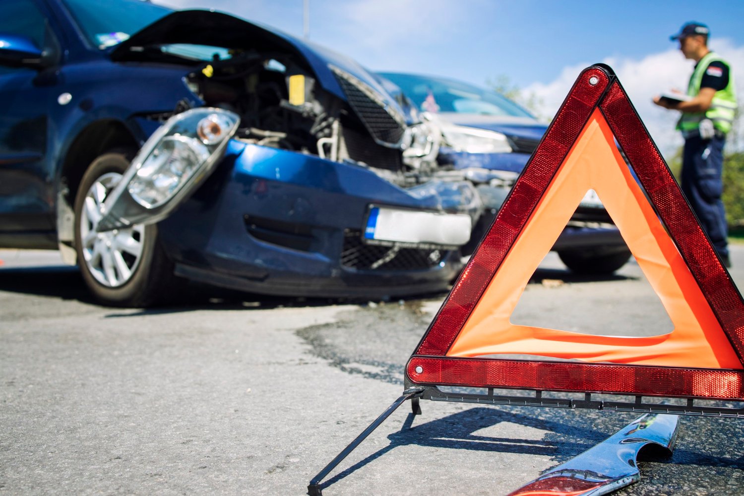 A reflective warning triangle marks a car accident scene with two heavily damaged vehicles and a police officer in the background.