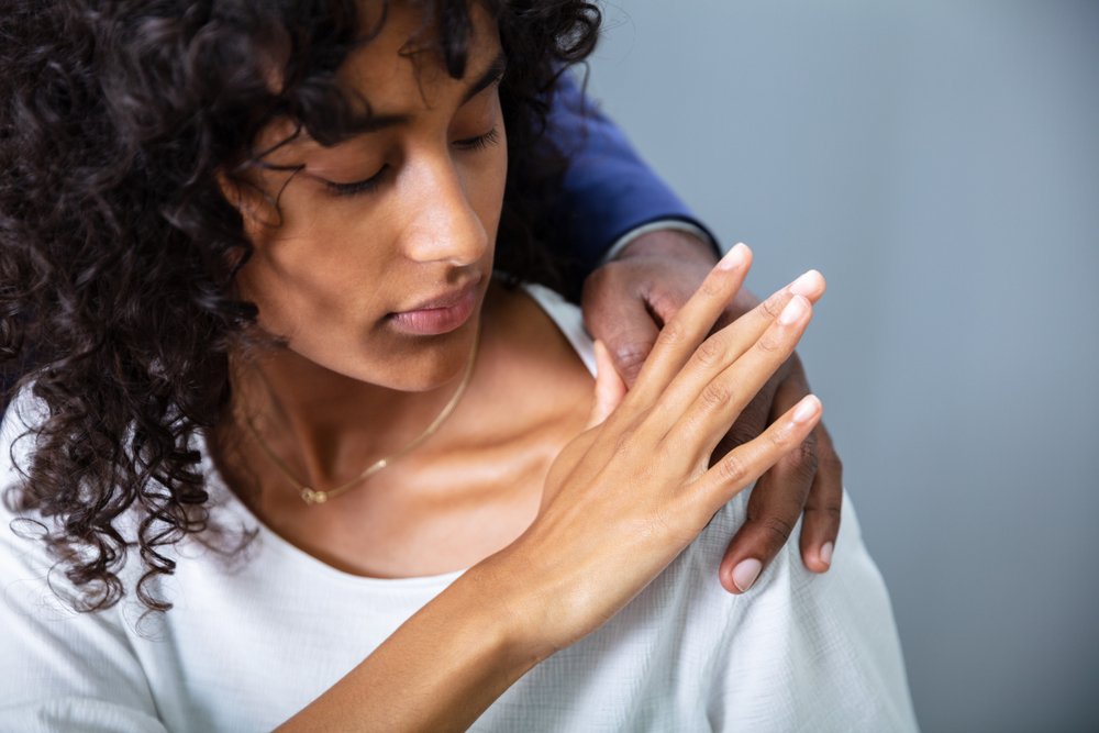 A woman with curly hair looks down with discomfort as a man's hand rests on her shoulder