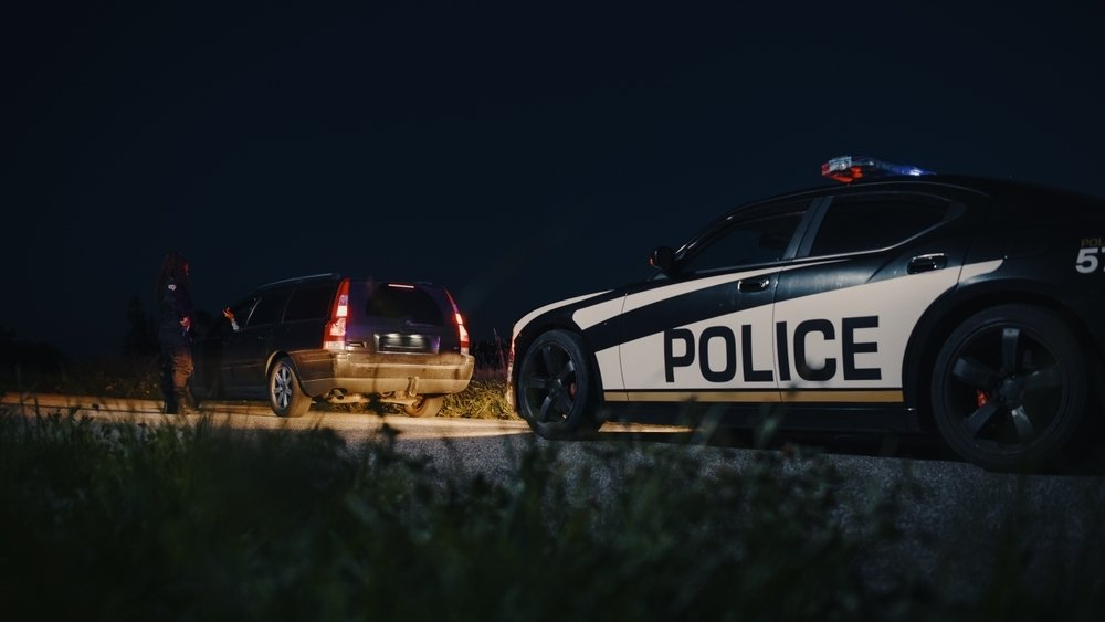 A police officer interacts with a driver during a nighttime traffic stop, with a police car, labeled POLICE, parked nearby.