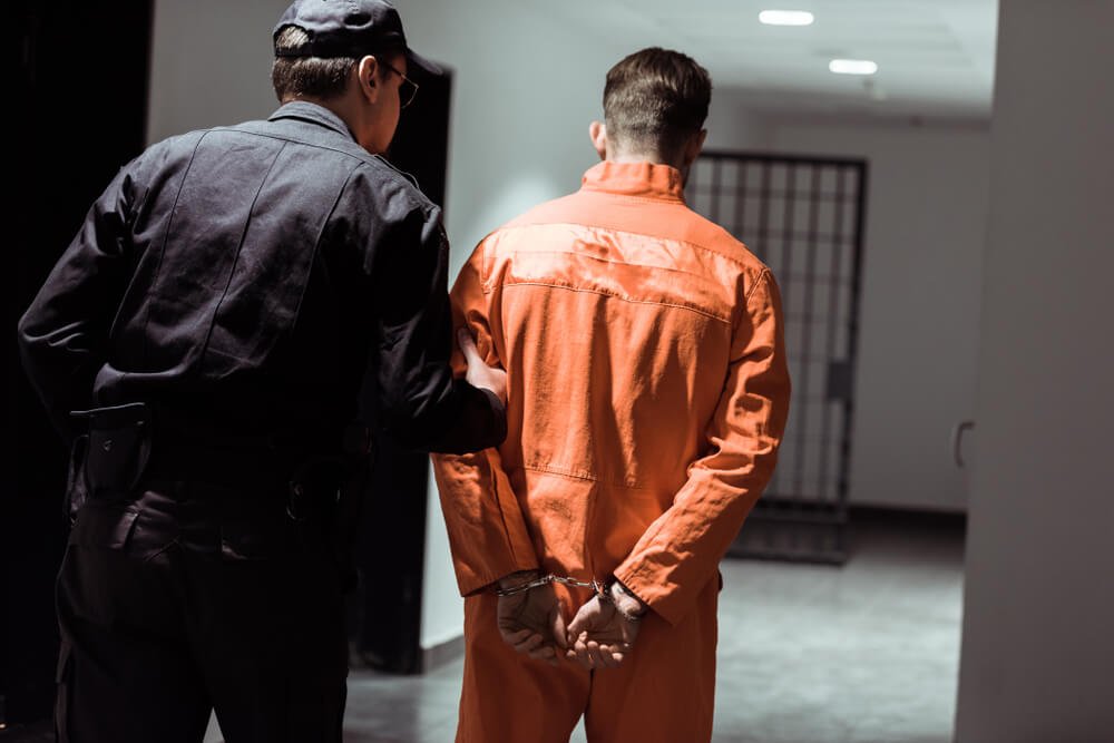 A law enforcement officer escorts a handcuffed person in an orange jumpsuit, representing a criminal defense scenario.