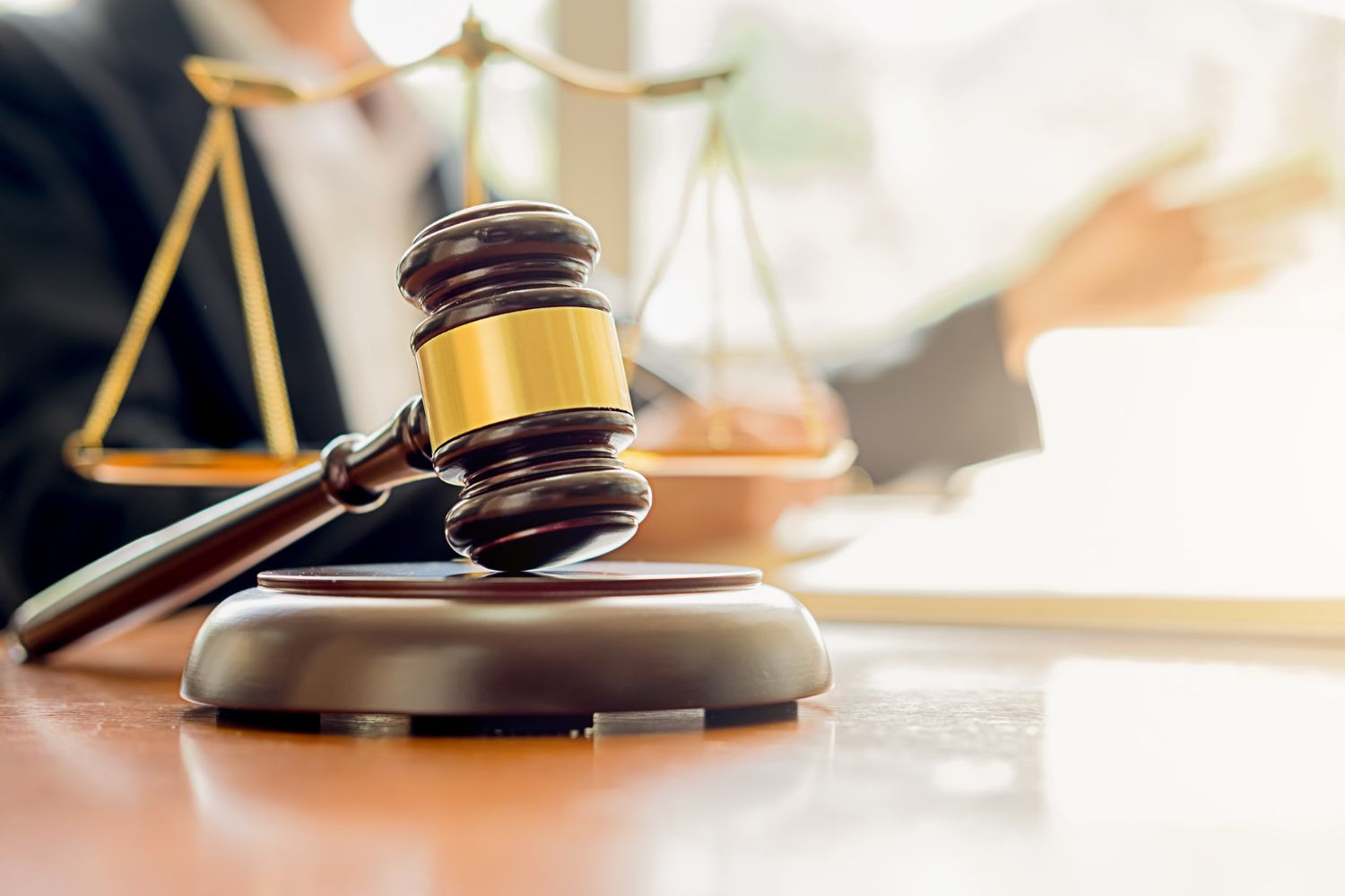 A close-up of a gavel on a table, with a blurred lawyer and scales of justice in the background, representing law.