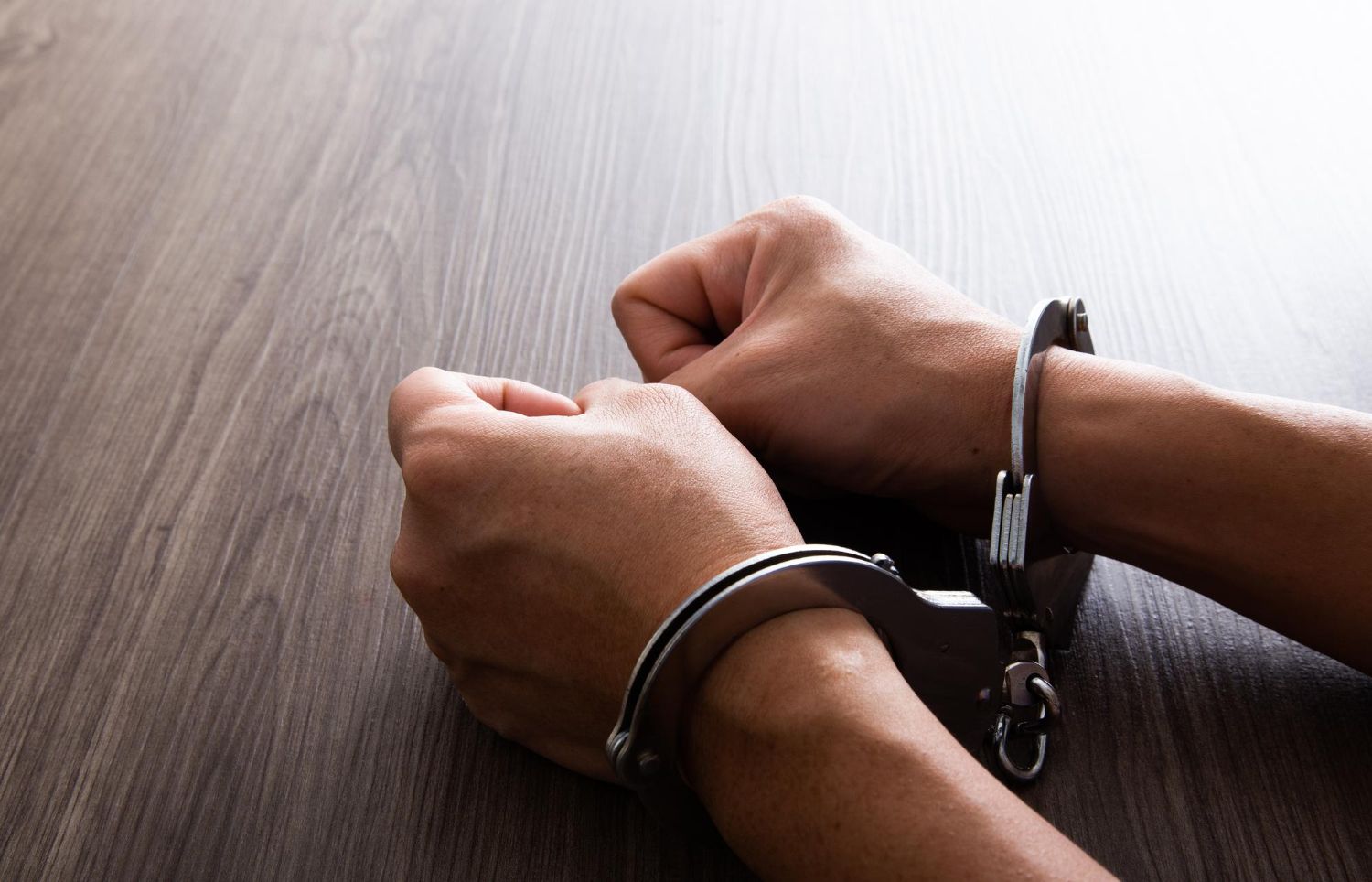 A close-up shows hands handcuffed, resting on a wooden table with visible grain