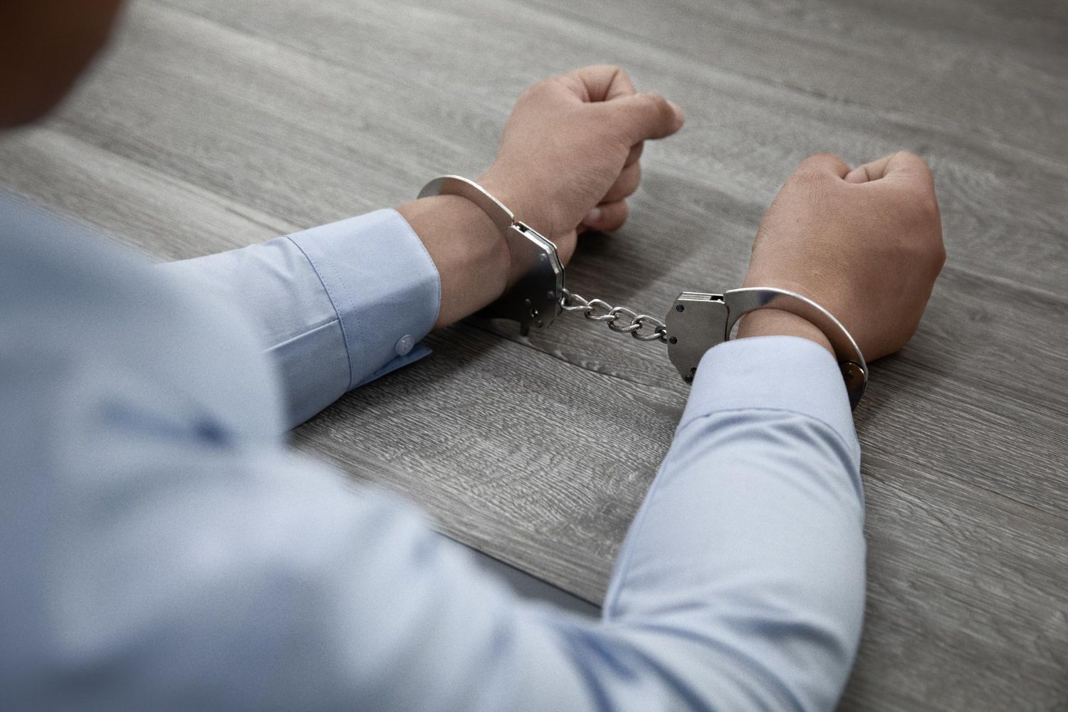 A person's hands, wearing a light blue dress shirt, are shown handcuffed and resting on a gray wooden table