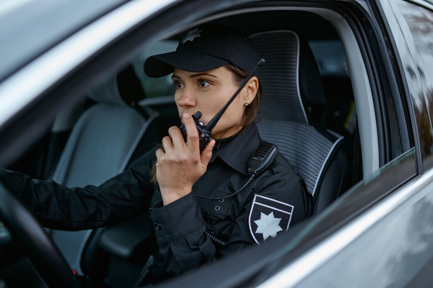 A female police officer in a black uniform and cap sits in a patrol car