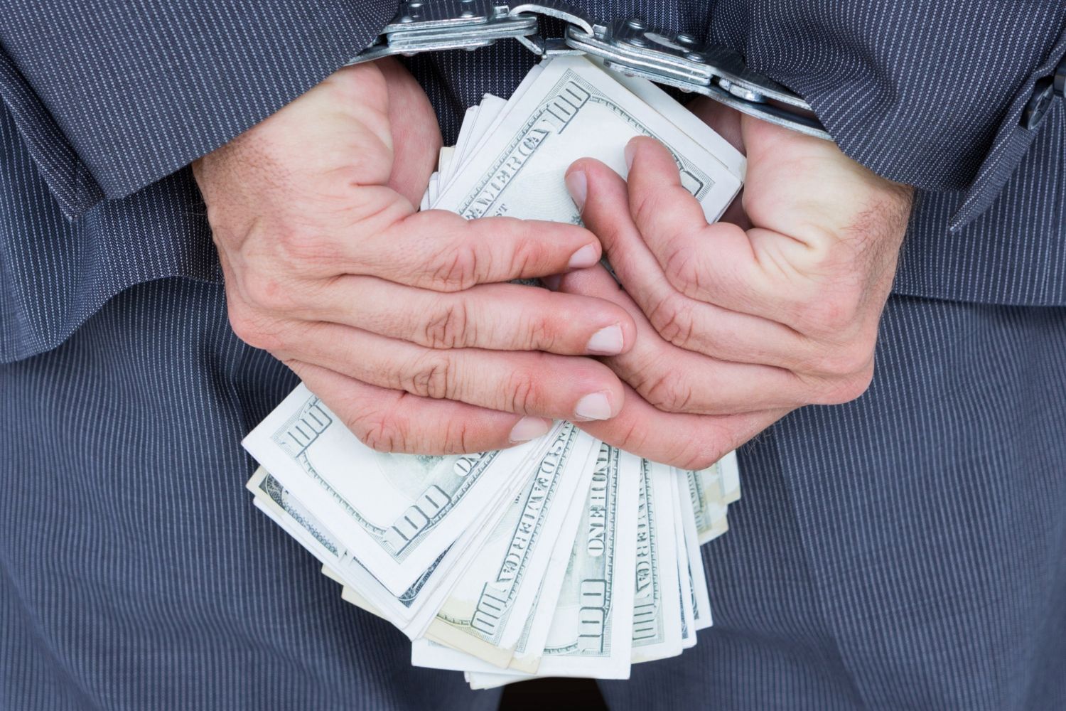 A person in a pinstriped suit has their cuffed hands, holding a stack of dollar bills, behind their back